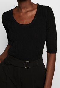 Musta ribattu toppi, jossa on viehättävä pääntie ja lyhyet hihat, yhdistettynä korkeavyötäröisiin housuihin, joissa on pyöreä hopeinen solkivyö.