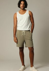 Witte tanktop met groene strepen, gedragen met olijfgroene textuurshorts met een elastische tailleband en trekkoord, aangevuld met witte instapschoenen.