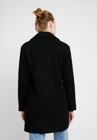 Manteau noir texturé avec col, manches longues et coupe droite. En tissu pelucheux, atteignant le milieu de la cuisse. Porté avec un jean bleu clair.