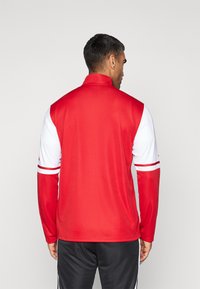 Rode en witte sportjack met een hoge kraag, lange mouwen en horizontale strepen op de mouwen, gemaakt van een gladde, lichte stof.