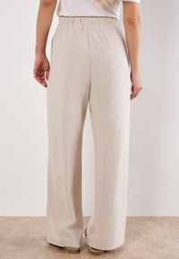 Vrouw in loose, high-waisted beige broeken met elastische tailleband, gezien van achteren, staande op een lichtbruine vloer.