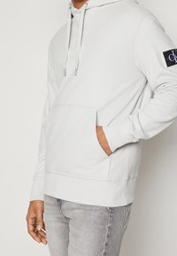 Ljust grå hoodie av mjukt tyg, med en känguruficka, justerbara dragsnören och en logotyp-patch på vänstra armen.