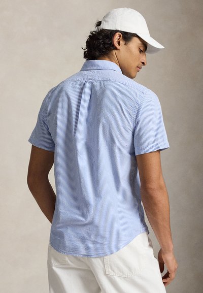 Homme portant une chemise à manches courtes rayée bleu clair et blanc, pantalon blanc et casquette blanche, vu de dos sur un fond uni.