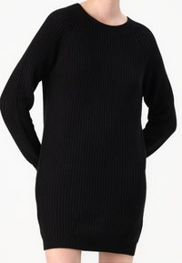 Robe pull en maille côtelée noire à manches longues portée par une personne se tenant les mains derrière le dos, sur fond blanc.