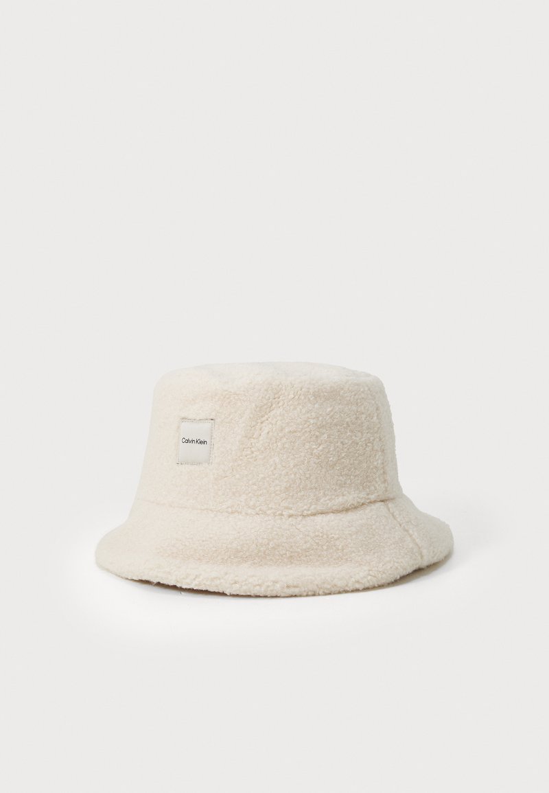 Klobouk s širokým okrajem vyrobený z měkkého krémového materiálu. Má texturovaný povrch a malou etiketou Calvin Klein po straně.