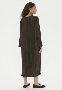 Largo vestido marrón con plisados, de manga larga, escote redondo y sutil dobladillo con volantes. Estilizado con zapatos planos negros.