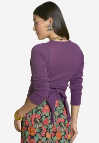 Purple knit sweater with long sleeves, tie-back detail at the waist, paired with a floral-patterned skirt featuring vibrant colors.