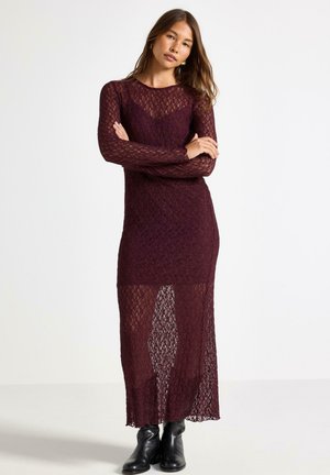Burgundy spetsklänning med långa ärmar, en figurnära silhuett och en genomskinlig, texturerad yta. Matchas med svarta ankelstövlar för kontrast.