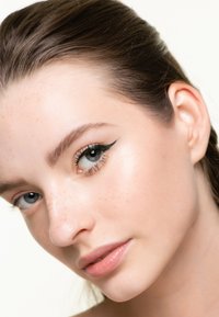 Primo piano di un volto con pelle chiara, lentiggini, sopracciglia definite e trucco occhi a cateye. Le labbra sono leggermente lucide, con una texture liscia.