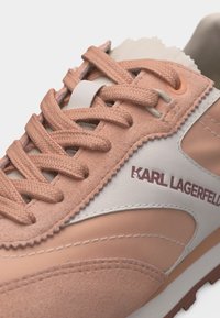 Κοντινό πλάνο αθλητικού παπουτσιού σε ροδακινί και λευκό χρώμα με υφασμένες κορδόνια, πάνελ από σουέτ και δέρμα, και το λογότυπο «KARL LAGERFELD» στην πλευρά.