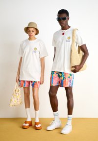 T-shirts brancas oversized, calções de banho florais coloridos, sapatos de plataforma laranja, bolsa de malha, chapéu bege claro, óculos de sol com tonalidade azul.