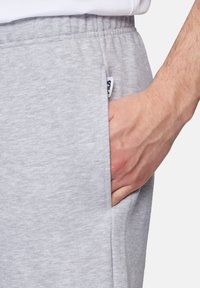 Pantalones de chándal gris claro con cinturilla elástica, tejido texturado, bolsillo lateral y una pequeña etiqueta de marca visible en el lado.