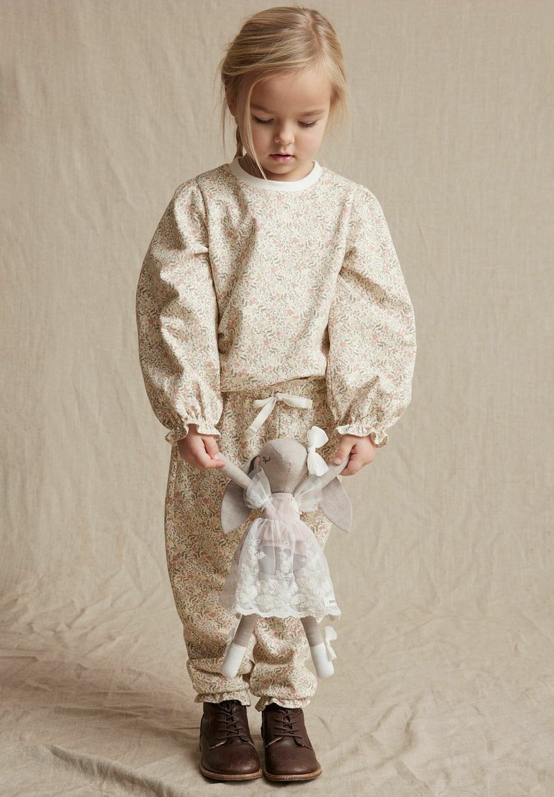 Krämfärgad loungewear-set i blommönster med pastellfärger, med rynkade ärmar och elastiska manschetter. Barnet håller en tygdocka.