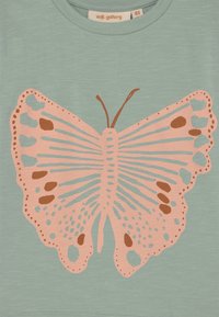 Jasnozielony materiał z dużą, stylizowaną grafiką motyla w różowym i brązowym kolorze, umieszczoną centralnie poniżej okrągłego dekoltu.
