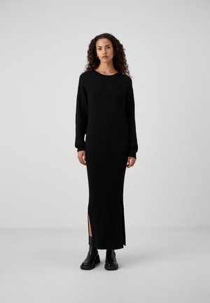 Long black ribbed dress with long sleeves, round neckline, and side slits. Model wears black ankle boots. Smooth texture and body-hugging fit.