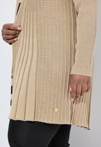 Cárdigan de punto acanalado beige con detalle plisado, llevado sobre pantalones negros, mostrado con dos manos ajustando la tela.