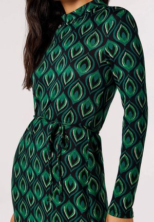 Femme portant une robe à manches longues verte et noire avec un motif abstrait de feuilles et une ceinture nouée à la taille.