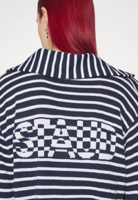 Marineblauw en witte gestreepte gebreide cardigan met een groot gebreid "STAUD" logo op de achterkant, voorzien van een brede kraag en een zachte textuur.