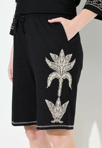 Pantaloni corti neri realizzati in un tessuto morbido, con un intricato ricamo bianco a forma di foglia sui lati e una vita elastica con lacci.