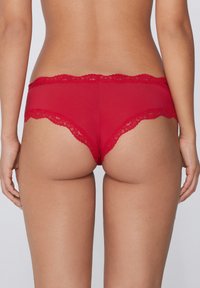 Intimo con pizzo rosso, caratterizzato da un design in tessuto trasparente, con bordi ondulati e una forma a vita bassa. Texture liscia, copertura minima.