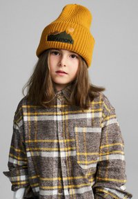 Musztardowa dzianinowa czapka typu beanie z prążkowaną fakturą i wyszywanym logo. Brązowa flanelowa koszula w kratę z żółtymi i białymi akcentami oraz kieszenią na piersi.