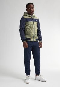 Giacca a vento verde e blu navy con texture trapuntata, chiusura con zip e polsini a coste, abbinata a pantaloni tuta blu navy e sneakers bianche.