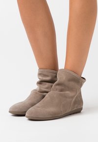 Botines de ante suave en color taupe con un diseño arrugado, llevados sobre piernas desnudas contra un fondo blanco liso.