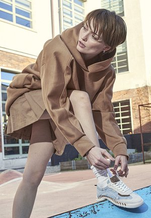 Beige oversized hoodie with loose sleeves, paired with white and blue sneakers featuring textured accents and laces, set against an outdoor background.