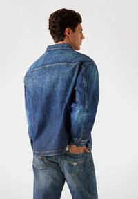 Denim jacket in medium blue with faded texture. Features a shirt collar, button closure, and two pockets on the back. Long sleeves.