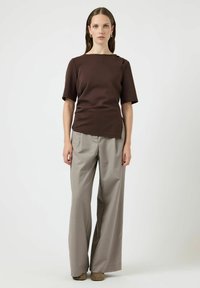 Maglietta marrone a maniche corte con collo alto e spacco laterale, abbinata a pantaloni a gamba larga grigio chiaro e stivali alla caviglia beige.