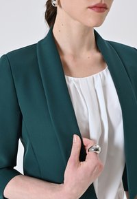 Blazer su misura teal con collo sciallato, indossato sopra una camicetta bianca plissettata. Presenta un anello d'argento alla mano, che mette in mostra una superficie liscia.
