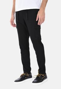 Pantaloni cargo neri con vestibilità slim, vita elasticizzata, tasche laterali e dettagli neri. Indossati con scarpe nere con suole bianche.