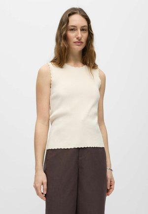 Jeune femme aux cheveux bruns ondulés, portant un top côtelé crème sans manches et un pantalon taille haute marron foncé sur fond blanc.