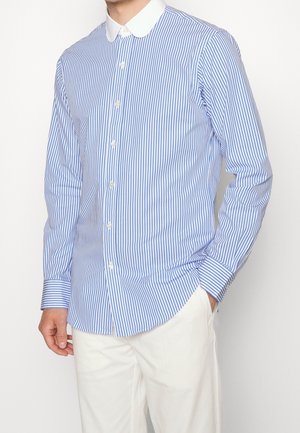 Hombre con camisa de manga larga a rayas verticales azules y blancas con cuello blanco y pantalones blancos, una mano en el bolsillo.