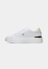 Sneaker bianche basse con suola spessa, logo dorato sul lato, linguetta beige sul tallone e dettagli metallici sul tallone.