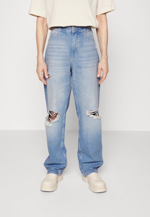 Light blue high-waist ripped jeans with large tears on both knees worn with cream chunky shoes and a cream short-sleeve top.