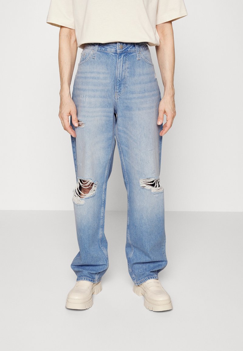 Svetle modré vysoké džíny s roztržením, které mají velké díry na obou kolenou, nošeny s krémovými robustními botami a krémovým tričkem s krátkým rukávem.