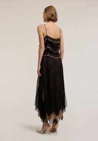 Vestito nero con design a balze sovrapposte sul corpetto, realizzato in materiale trasparente con dettagli in pizzo dorato sull'orlo, abbinato a décolleté nude.