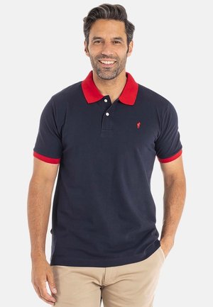 Homme souriant, portant un polo marine avec un col et des manches rouges, un pantalon beige, se tenant devant un fond clair.