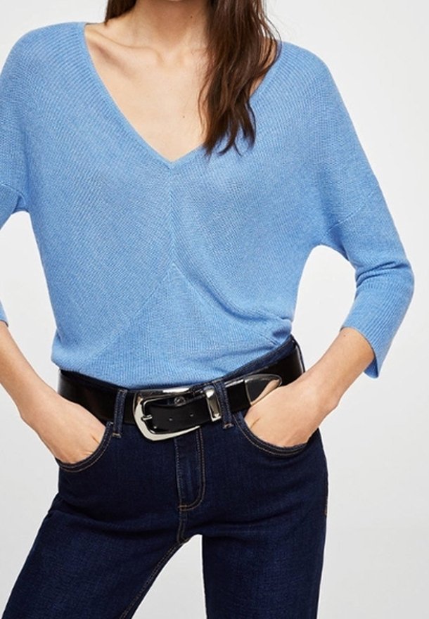 Femme portant un pull bleu clair à col en V avec des manches trois-quarts, un jean bleu foncé et une ceinture noire avec une boucle argentée.