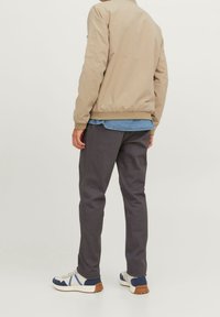 Ljusbeige bomberjacka, elastisk nederkant, matchad med mörkgrå byxor; modellen bär en denimskjorta under, vita sneakers med blå detaljer.