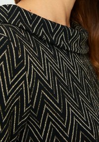 Černý svetr s vysokým límcem a béžovým cik-cak vzorem, který nosí osoba s hnědými vlasy viditelnými na okraji límce.