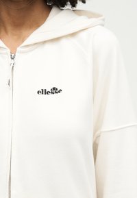 Κοντινό πλάνο ατόμου που φοράει λευκή μπλούζα με φερμουάρ και κουκούλα, με μαύρο λογότυπο ellesse στο αριστερό μέρος του στήθους.