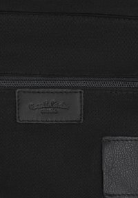 Tejido negro con una cremallera horizontal sobre una etiqueta de cuero cosida que dice "Guest Leather studio" en el borde texturizado de un bolsillo de cuero.