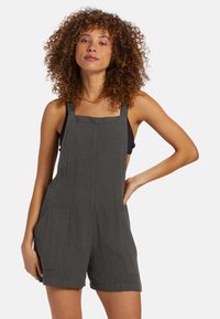 Salopette di cotone grigio scuro con bretelle regolabili, tessuto strutturato, due tasche frontali e vestibilità comoda, abbinata a una maglia nera.