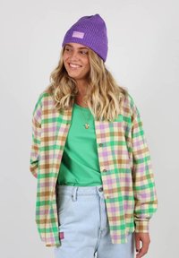Chemise en flanelle à carreaux verts, violets et bruns avec des manches longues, boutons en bois et un T-shirt vert en dessous. Portée avec un bonnet violet.