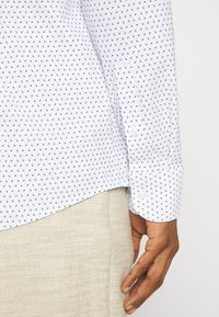 Witte shirt met kleine blauwe patronen, lange mouwen, met knoopmanchetten. De stof lijkt glad met een lichte glans; de zoom is afgerond. Beige broek is zichtbaar.