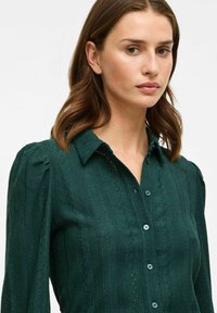 Blusa button-up verde scuro con tessuto testurizzato caratterizzato da strisce verticali e cuciture decorative lungo il davanti. Collo e maniche lunghe.