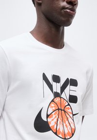 Άνδρας με σκουρόχρωμο δέρμα, που φοράει λευκό μπλουζάκι Nike με μαύρο λογότυπο και πορτοκαλί γραφικό με θέμα το μπάσκετ στο μπροστινό μέρος.