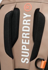Beige tyg med svarta nätaccent, som har kraftig vit "SUPERDRY"-logotyp och en orange detalj i siden.
